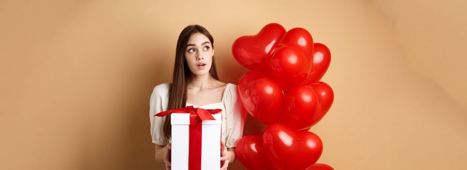Valentines day. Pensive cute girl guessing who made her gift, holding present and looking curious at upper left corner, standing near romantic heart balloons, beige background.