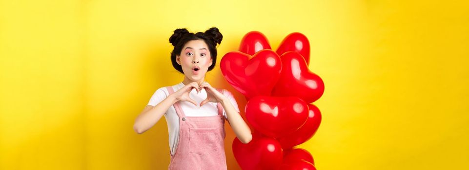 Surprised asian woman looking in awe, showing heart gesture, making love confession on valentines day, standing near romantic red balloons over yellow background.