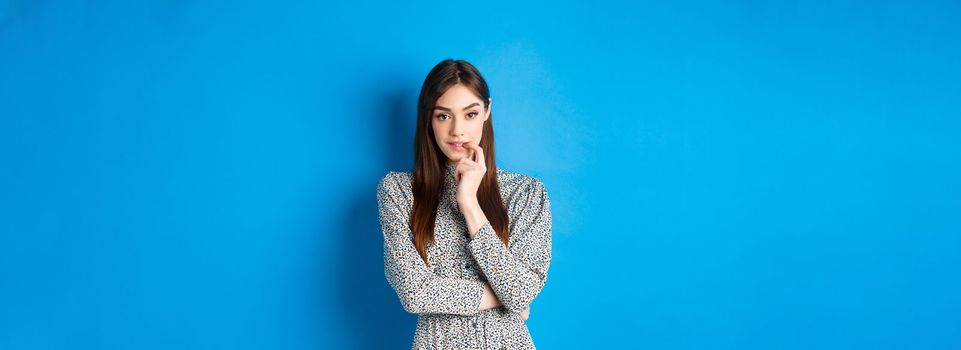 Sensual woman in dress biting finger and looking pensive at camera, standing dreamy against blue background.