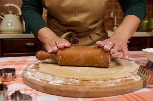 Cropped view hands of housewife in beige chef apron, baker confectioner rolls out dough, using rolling pin, cooking homemade gingerbread cookies, according to traditional family recipe in the kitchen