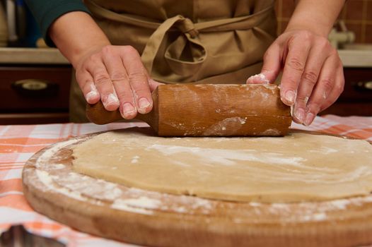Details: Housewife, woman or chef confectioners hands using rolling pin, roll out the dough on a floured wooden board. Baking concept. Making pizza, bread, festive homemade pastries. Cookery. Culinary