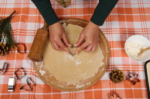 View directly above a female chef confectioner hands cutting a heart shaped mold on a gingerbread dough, making homemade pastries for Christmas.