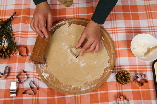 Details: hands of a housewife carving molds from the dough, preparing homemade gingerbread cookies for Christmas dinner. Holiday bakery. Cookie cutters, pine cones and fir-tree branch on the table