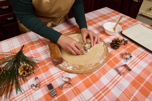 Cropped view. Housewife in green shirt and beige chef apron, stands by a kitchen table and carves cookies from gingerbread dough. Cookie cutters and branch of fir-tree and pine cones lie on the table