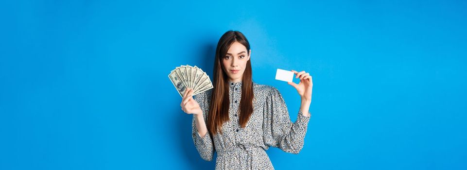 Shopping. Elegant sassy woman with dollar bills money and plastic credit card, looking confident at camera, blue background.