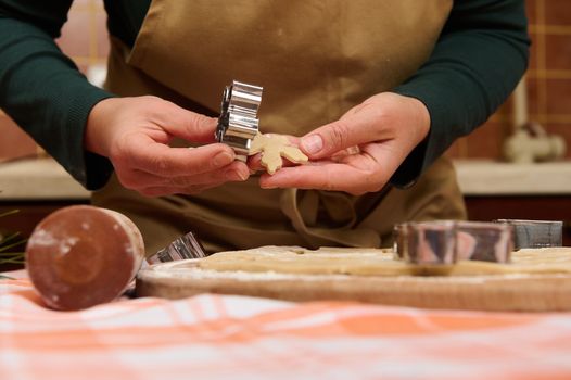 Details: hands of housewife confectioner, baker, pastry chef in green shirt and beige apron, removing cut snowflake shaped mold from cookie cutter while making Christmas gingerbread biscuits, pastries