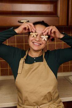Cheerful housewife in green shirt and beige apron, holding two snowflake shaped gingerbread dough near her eyes and smiling a beautiful toothy smile, while baking christmas cookies in the home kitchen