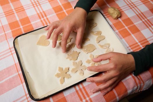 Top view hands of a housewife chef pastry, female confectioner putting cut molds of raw dough on the baking sheet, before placing gingerbread cookies in the hot oven. Christmas culinary and bakery