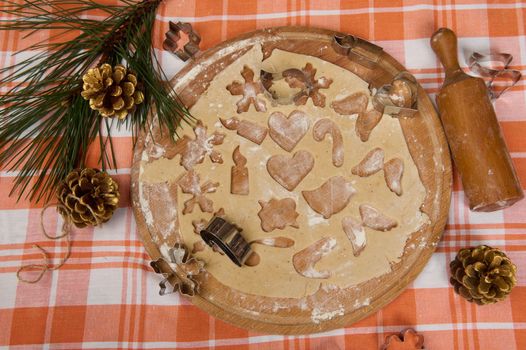 Top view of rolled out gingerbread dough on a wooden board sprinkle with flour, on the table with red plaid tablecloth and pine branch, cones and rolling pin. Making festive pastries. Merry Christmas