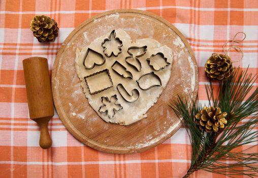 View from above of cookie cutters on the gingerbread dough, rolled out on a wooden board, next to a rolling pin and fir-tree branch with golden pine cones. Merry Christmas concept. Festive baking