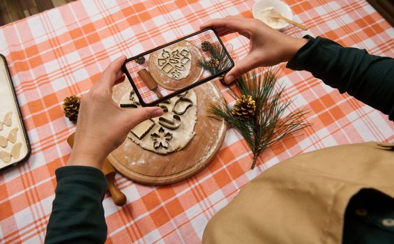 View from above to housewife's hands holding smartphone in live view mode, photographing rolled out gingerbread dough and cookie cutters, on the kitchen table. Merry Christmas concept. Festive cookery