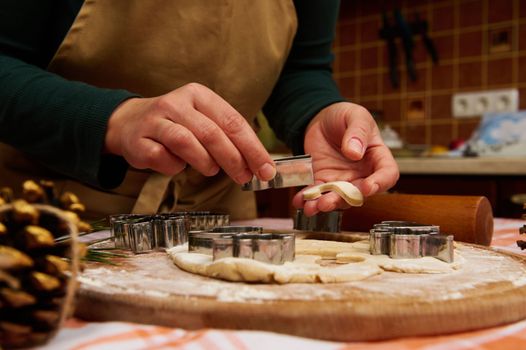 Details: hands of housewife confectioner, baker, pastry chef in green shirt and beige apron, removing cut candy cane shaped mold from cookie cutter while making Christmas gingerbread biscuits