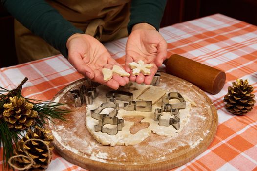 Carved snowflake and angel wings made of ginger dough in the hands of a housewife, pastry chef, preparing gingerbread cookies for Christmas. On the table are golden pine cones on the fir-tree branch