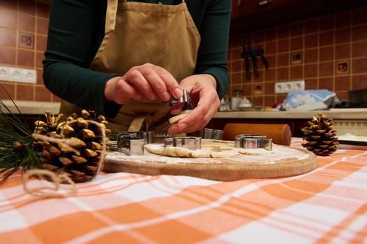 Close-up unrecognizable woman housewife, a confectioner amateur wearing green shirt and beige chef apron, cutting gingerbread dough with cookie cutters on a table. Christmas atmosphere in the kitchen