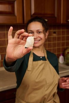 Selective focus. Carved bird from gingerbread dough in the hands of a blurred charming multi-ethnic woman housewife confectioner in beige apron, smiling a cheerful toothy smile looking at the camera