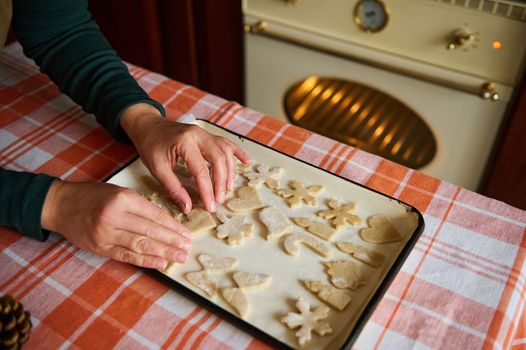 Close-up woman's hands put gingerbread cookies on baking sheet, standing by a table against a heating oven. Christmas, New Year celebration traditions. Traditional festive food making, family culinary