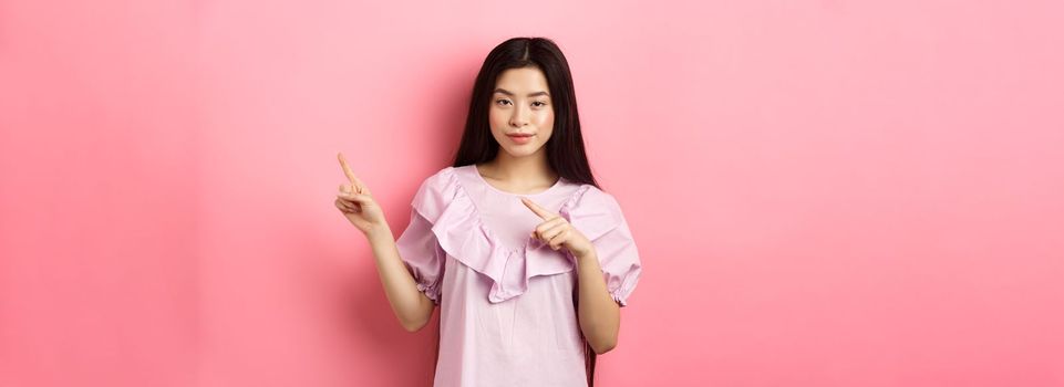 Cheeky asian girl showing interesting advertisement, pointing fingers right at logo and smiling cunning, hinting on good deal, pink background.