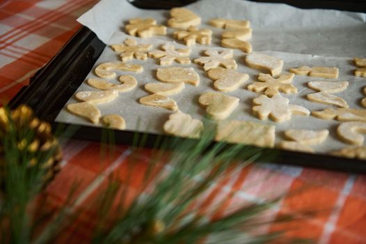 View through blurred fir-tree needles and golden pine cones, to a baking sheet with carved molds of gingerbread dough before baking in the oven. Christmas background. Still life. Festive pastries