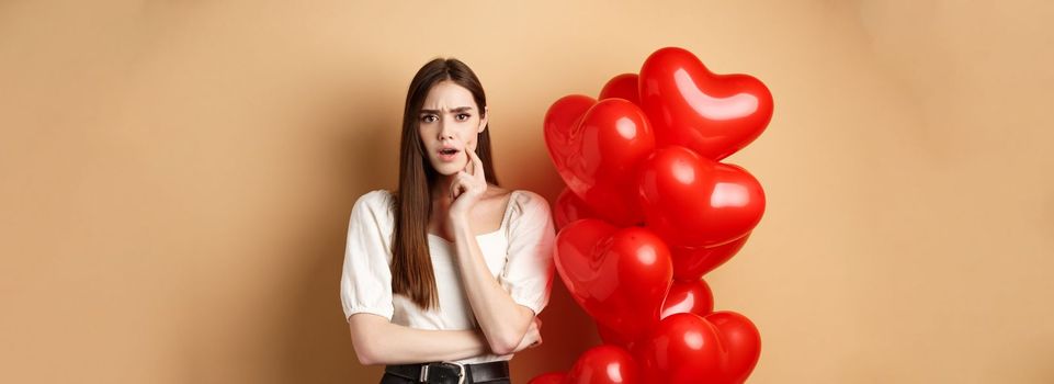 Valentines day and love concept. Confused and shocked girl staring displeased at camera, standing near romantic red hearts balloons on beige background.