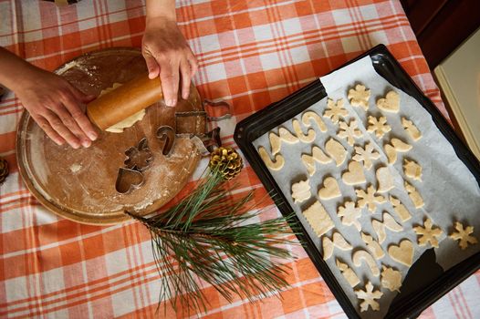 Top view. Selective focus. Housewife's hands roll out dough with a rolling pin, a baking sheet with carved molds of gingerbread dough on the table. Preparing homemade Christmas pastries in the kitchen