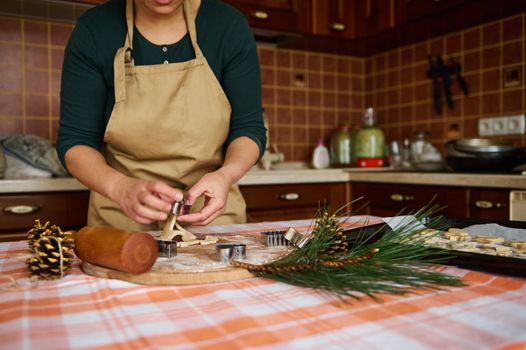 A woman housewife, wearing a green shirt and a beige chef's apron, using cookie cutters, preparing gingerbread cookies of various shapes, for Christmas in the kitchen at home. Pine cones as ornament