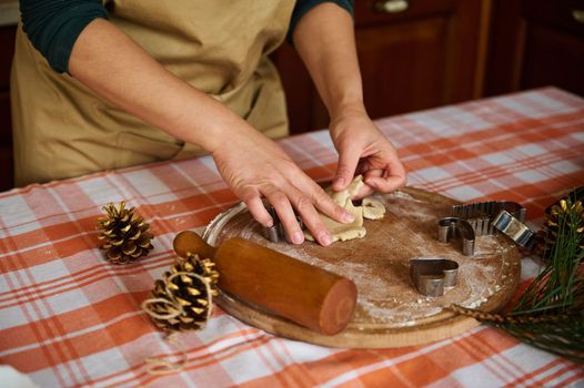 Close-up of a woman housewife, wearing a beige chef's apron, preparing homemade gingerbread cookies for Christmas in the kitchen at home. Female confectioner using cookie cutter, making biscuits