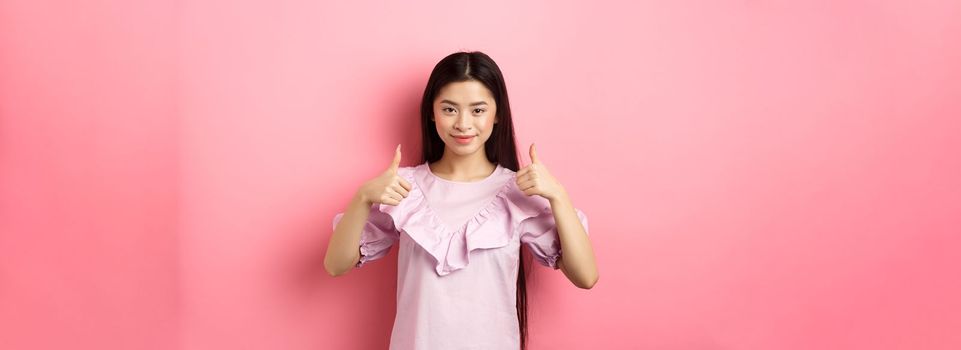 Well done. Smiling asian girl in dress showing thumbs up, like and approve product, praise great deal, standing against pink background.
