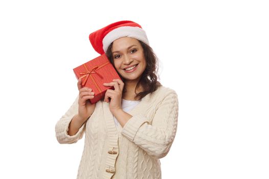 Multi-ethnic middle-aged pretty woman wearing Santa hat, holding a Christmas gift box near her face, smiling a cheerful toothy smile looking at camera, isolated on white background, copy ad space