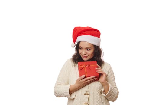 Beautiful African American woman wearing Santa hat and beige sweater, holding Christmas present over white background. Boxing Day. Time to open gift boxes. New Year's preparations. Christmastime