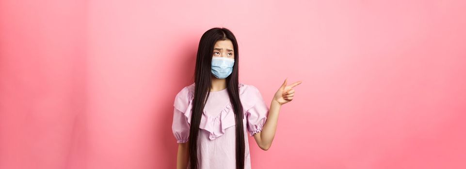 Coronavirus, quarantine and lifestyle concept. Sad and gloomy asian woman in medical mask pointing, looking left upset, showing unfair thing, standing on pink background.