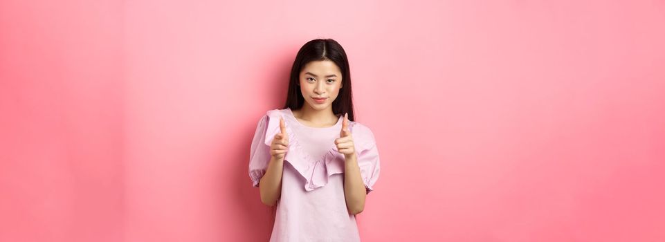 Sassy asian woman pointing at camera, smiling and inviting you, beckon or praise person, standing on pink background.