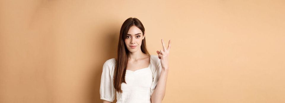 Attractive young woman show fingers number two, smiling and looking confident, standing on beige background.