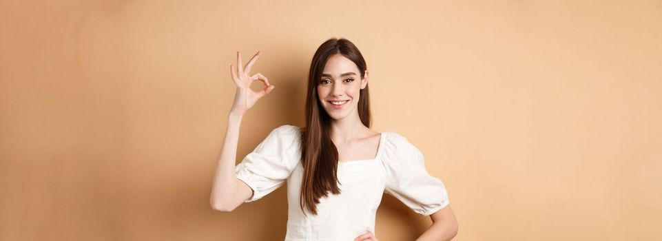 Very good. Smiling cute woman in white blouse showing okay sign in approval, like and praise excellent choice, standing satisfied on beige background.