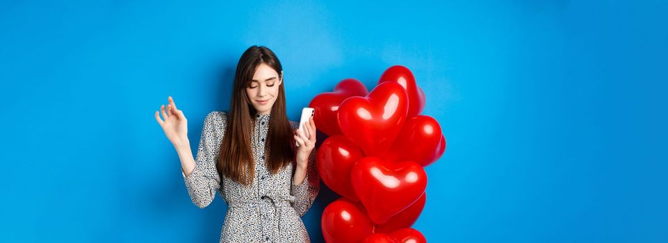 Portrait of attractive young woman holding smartphone and dancing near Valentines red balloons, standing on blue background.