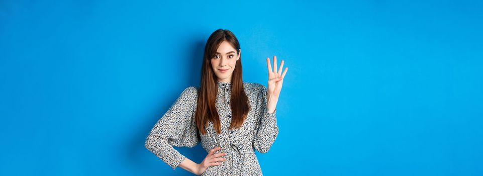 Young beautiful girl in dress with natural long hair, showing number four with fingers and smiling, standing against blue background.