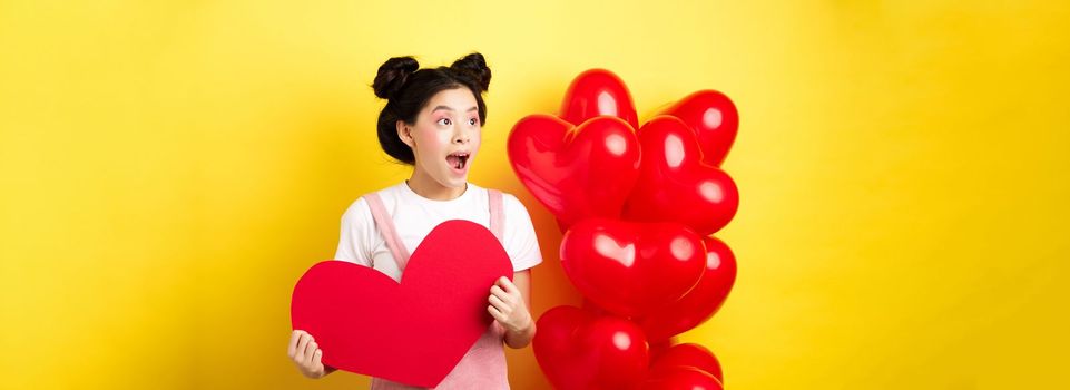 Happy Valentines day. Excited and surprised asian girl checking out romantic offer, looking left cheerful, showing big red heart card, yellow background.