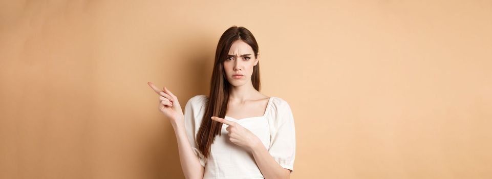 Angry young woman frowning, pointing fingers right and complaining, disappointed with logo, standing upset on beige background.