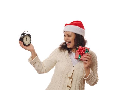 Happy woman wearing Santa hat, holding a cute gift box for Christmas, rejoices looking at the alarm clock in her hands, with five to midnight on the dial. Looking forward at upcoming winter holidays