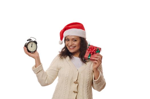 Delightful woman wearing Santa hat, holding a happy Xmas gift box with surprise, and black retro alarm clock showing five minutes to midnight on dial, smiles looking at camera, over white background