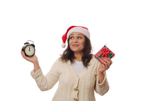 Mysterious multi-ethnic woman in Santa hat, holding a Christmas or New Year gift box and retro alarm clock showing five minutes to midnight, thoughtfully looking up at copy space on white background