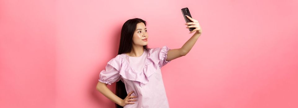 Stylish asian girl taking selfie and smiling, posing for social media photo, standing in dress against pink background.