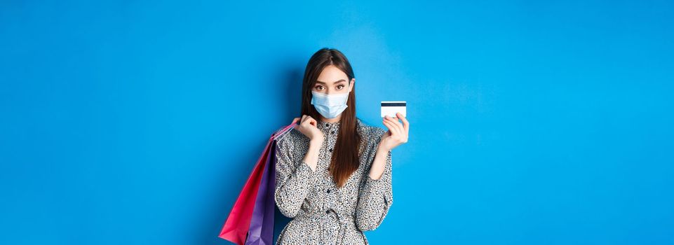 Covid-19, pandemic and lifestyle concept. Stylish woman wearing medical mask on shopping as preventive measure from coronavirus, showing plastic credit card and holding bags, blue background.