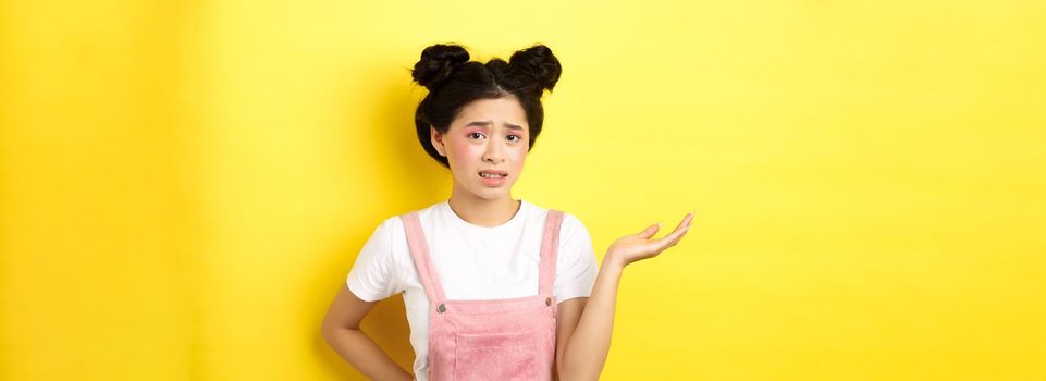 Confused teen asian girl with bright makeup and summer clothes, raising hand up and frowning puzzled, cant understand, yellow background.