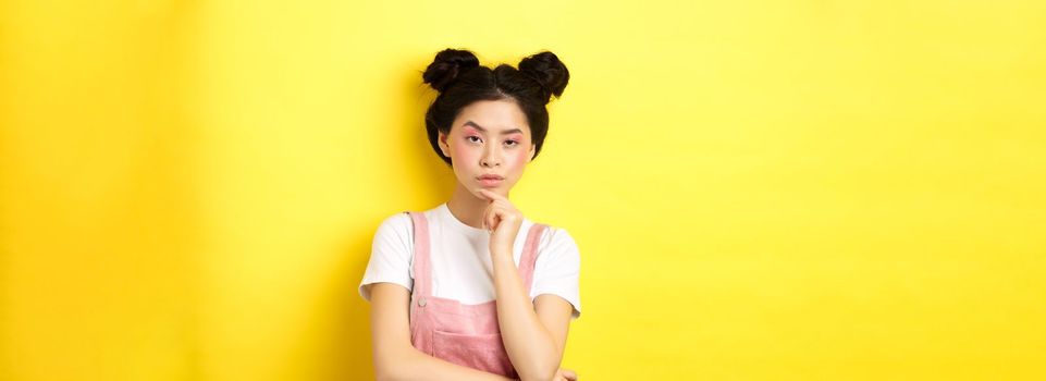 Suspicious asian girl looking at camera and thinking, stare with disbelief, standing with makeup on yellow background.