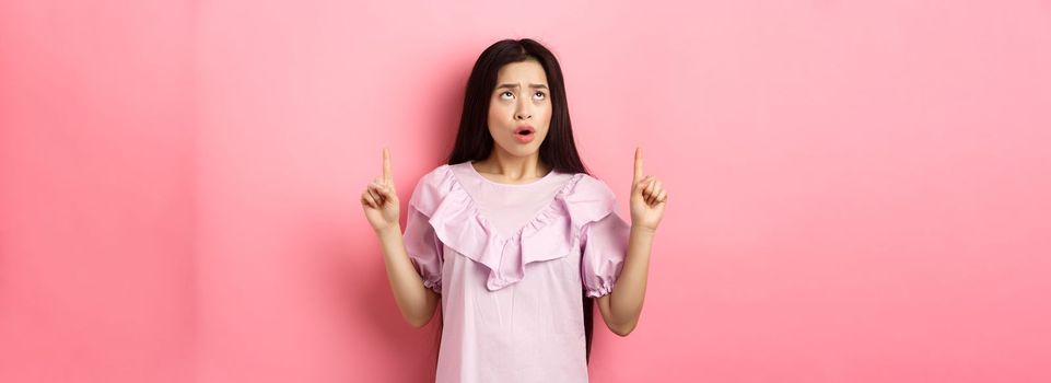 Concerned and sad asian girl frowning, looking and pointing fingers up at bad news, standing against pink background.