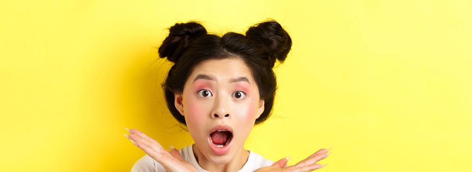 Close-up of excited asian woman with glamour makeup and hairstyle, screaming of amazement, open mouth and look at camera in awe, yellow background.