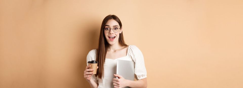 Excited woman in glasses drinking coffee from takeout cup, holding laptop and smiling happy, standing on beige background.