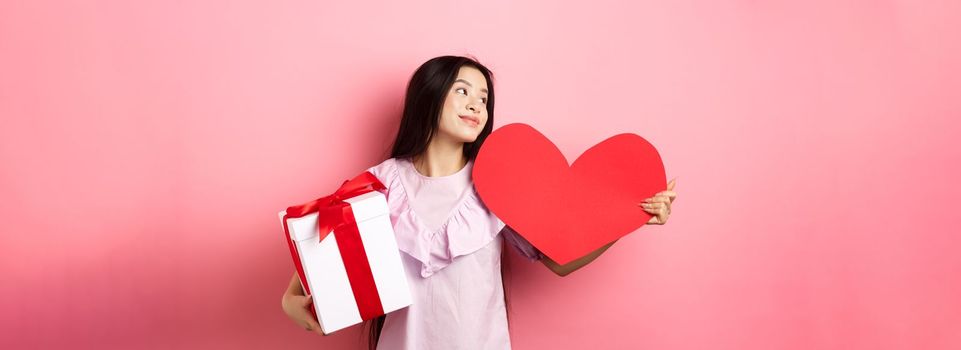 Valentines day concept. Romantic girl holding lover gifts, big red heart card and box with present, looking aside with dreamy look, falling in love, smiling tenderly, pink background.