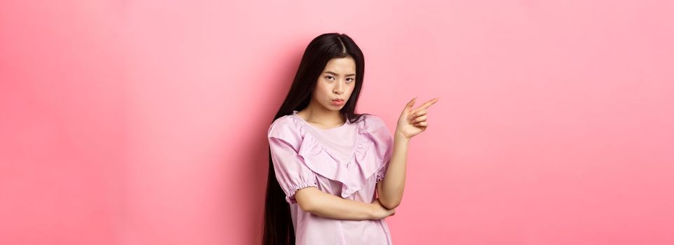 Sad and grumpy asian girl sulking, pointing left and look at camera jealous, want something, complaining and showing desired thing, standing on pink background.