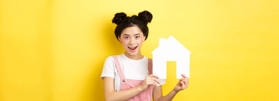 Real estate and family concept. Cute asian woman with bright makeup and stylish hairbuns, showing paper house cutout, smiling determined, yellow background.